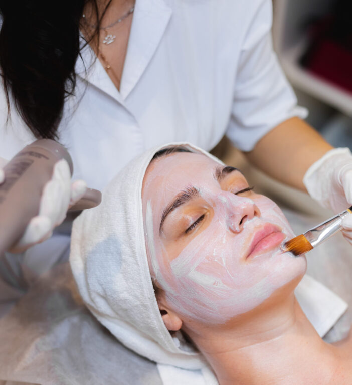 Beautician with a brush applies a white moisturizing mask to the face of a young girl client in a spa beauty salon